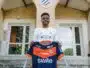 Young man stands on concrete steps outside a training center, holding a blue and orange football jersey toward the camera.