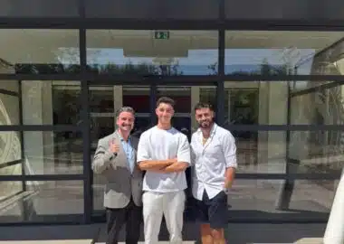 Three men stand outside a glass-front building, smiling for the camera; left man in a gray blazer gives a thumbs up, middle in white outfit, right in white shirt and shorts.