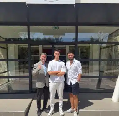 Three men stand outside a glass-front building, smiling for the camera; left man in a gray blazer gives a thumbs up, middle in white outfit, right in white shirt and shorts.