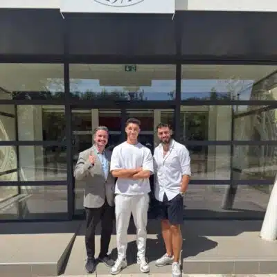 Three men stand outside a glass-front building, smiling for the camera; left man in a gray blazer gives a thumbs up, middle in white outfit, right in white shirt and shorts.