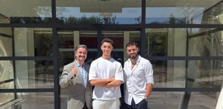 Three men stand outside a glass-front building, smiling for the camera; left man in a gray blazer gives a thumbs up, middle in white outfit, right in white shirt and shorts.
