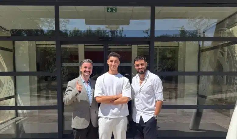 Three men stand outside a glass-front building, smiling for the camera; left man in a gray blazer gives a thumbs up, middle in white outfit, right in white shirt and shorts.