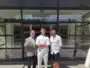 Three men stand outside a glass-front building, smiling for the camera; left man in a gray blazer gives a thumbs up, middle in white outfit, right in white shirt and shorts.