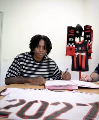 Screenshot | AllezPaillade.com Person signing documents at a table with a Flamengo jersey and scarves displayed in the background, suggesting a contract signing.