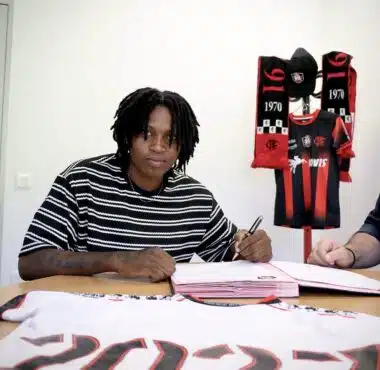 Person signing documents at a table with a Flamengo jersey and scarves displayed in the background, suggesting a contract signing.