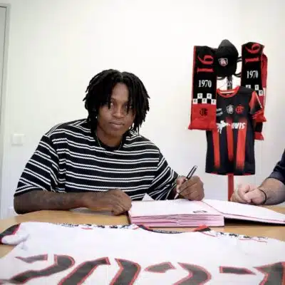 Person signing documents at a table with a Flamengo jersey and scarves displayed in the background, suggesting a contract signing.