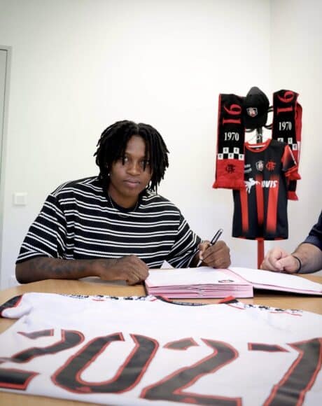 Person signing documents at a table with a Flamengo jersey and scarves displayed in the background, suggesting a contract signing.