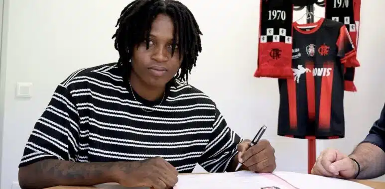 Screenshot | AllezPaillade.com Person signing documents at a table with a Flamengo jersey and scarves displayed in the background, suggesting a contract signing.