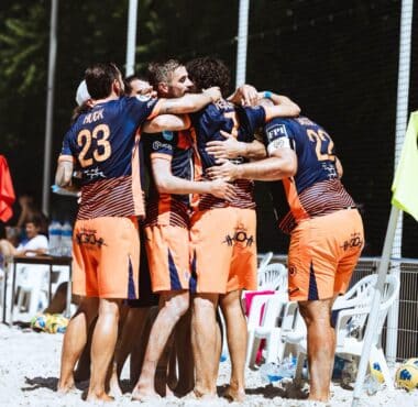 Beach volleyball team in blue-and-orange uniforms huddle together in celebration on sandy court near fencing and chairs.