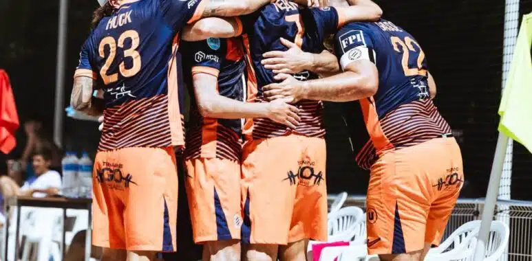 Beach volleyball team in blue-and-orange uniforms huddle together in celebration on sandy court near fencing and chairs.