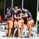 Beach volleyball team in blue-and-orange uniforms huddle together in celebration on sandy court near fencing and chairs.