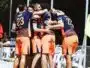 Beach volleyball team in blue-and-orange uniforms huddle together in celebration on sandy court near fencing and chairs.