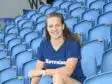 Smiling woman sitting in blue stadium seats, wearing a navy T-shirt with 'Havraise' logo.