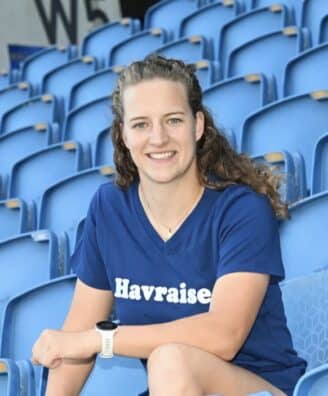 Smiling woman sitting in blue stadium seats, wearing a navy T-shirt with 'Havraise' logo.