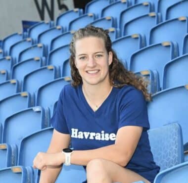 Smiling woman sitting in blue stadium seats, wearing a navy T-shirt with 'Havraise' logo.