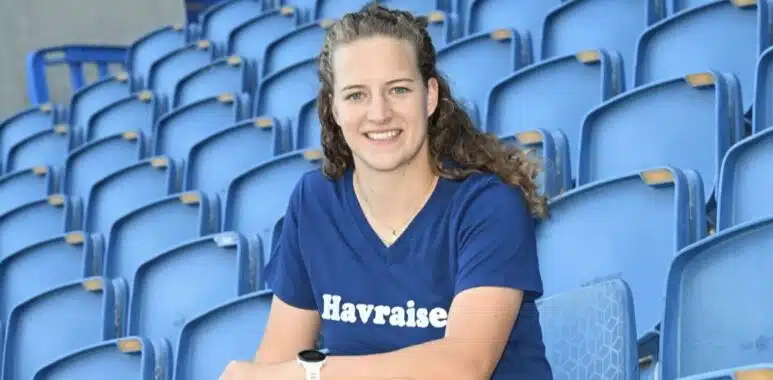 Screenshot | AllezPaillade.com Smiling woman sitting in blue stadium seats, wearing a navy T-shirt with 'Havraise' logo.