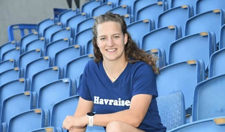 Smiling woman sitting in blue stadium seats, wearing a navy T-shirt with 'Havraise' logo.