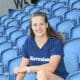 Smiling woman sitting in blue stadium seats, wearing a navy T-shirt with 'Havraise' logo.