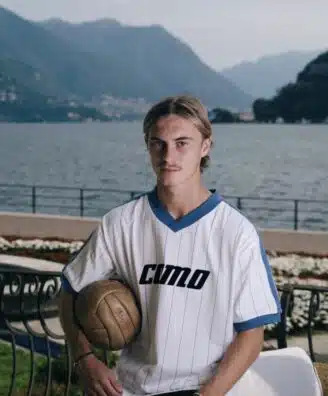 Young man in a white pinstriped soccer jersey holds a ball, posing by a lakeside with mountains in the background
