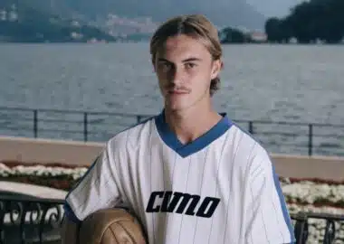 Screenshot | AllezPaillade.com Young man in a white pinstriped soccer jersey holds a ball, posing by a lakeside with mountains in the background