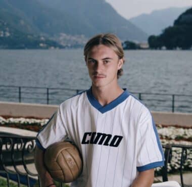 Young man in a white pinstriped soccer jersey holds a ball, posing by a lakeside with mountains in the background