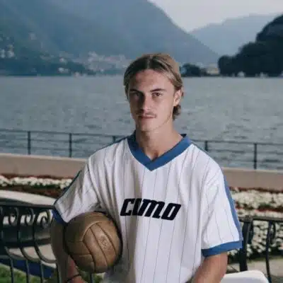 Young man in a white pinstriped soccer jersey holds a ball, posing by a lakeside with mountains in the background
