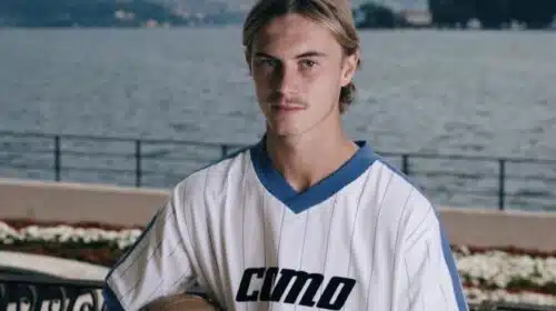 Young man in a white pinstriped soccer jersey holds a ball, posing by a lakeside with mountains in the background