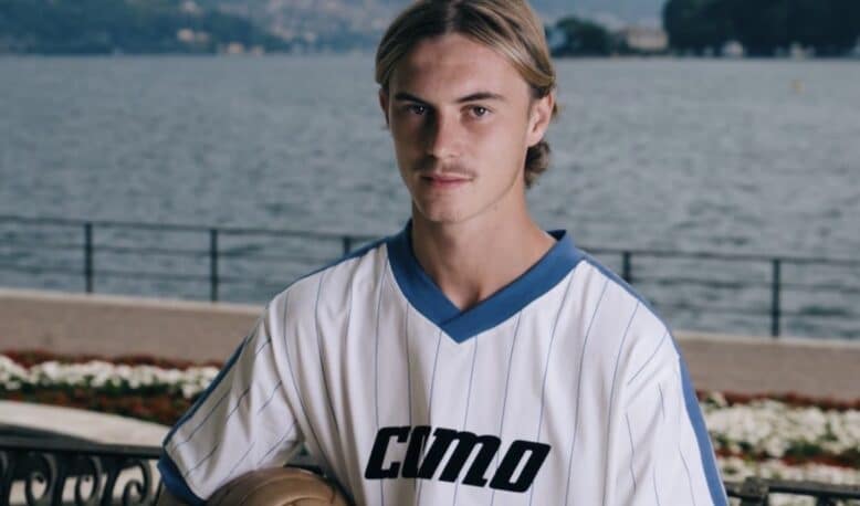 Screenshot | AllezPaillade.com Young man in a white pinstriped soccer jersey holds a ball, posing by a lakeside with mountains in the background