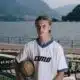 Young man in a white pinstriped soccer jersey holds a ball, posing by a lakeside with mountains in the background