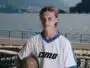 Young man in a white pinstriped soccer jersey holds a ball, posing by a lakeside with mountains in the background