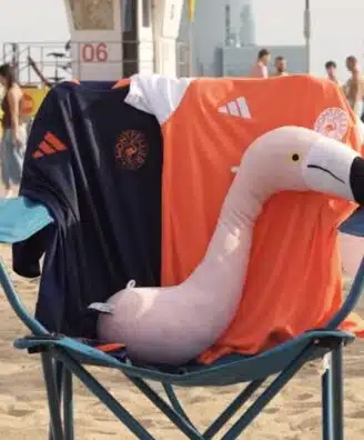 Pink inflatable flamingo resting on a blue beach chair, draped with dark blue and orange towels bearing logos.