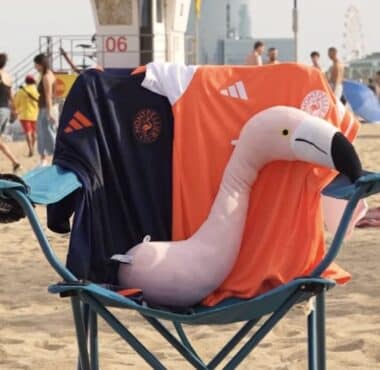Pink inflatable flamingo resting on a blue beach chair, draped with dark blue and orange towels bearing logos.