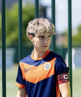 Young boy in an orange and navy soccer jersey stands behind a green metal fence, wearing a captain's armband.