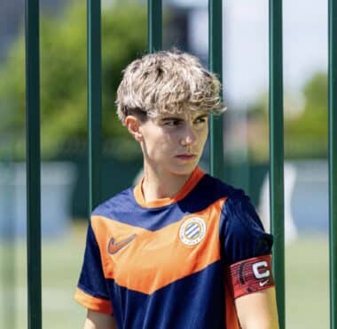 Young boy in an orange and navy soccer jersey stands behind a green metal fence, wearing a captain's armband.