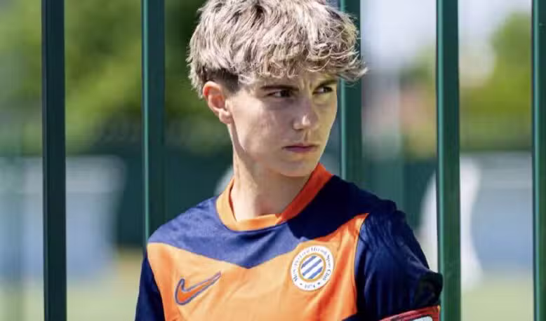 Young boy in an orange and navy soccer jersey stands behind a green metal fence, wearing a captain's armband.