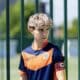 Young boy in an orange and navy soccer jersey stands behind a green metal fence, wearing a captain's armband.