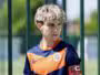 Young boy in an orange and navy soccer jersey stands behind a green metal fence, wearing a captain's armband.