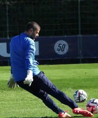 Soccer goalkeeper in blue jersey and white gloves kicking multiple balls on a green field.