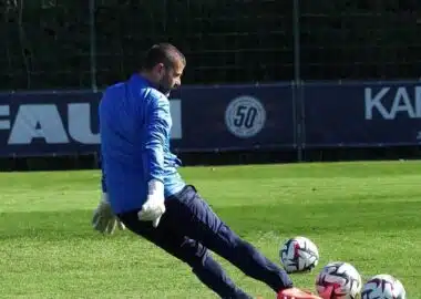 Soccer goalkeeper in blue jersey and white gloves kicking multiple balls on a green field.