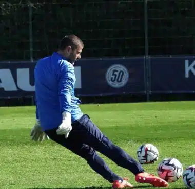 Soccer goalkeeper in blue jersey and white gloves kicking multiple balls on a green field.