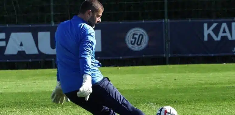 Soccer goalkeeper in blue jersey and white gloves kicking multiple balls on a green field.
