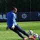 Soccer goalkeeper in blue jersey and white gloves kicking multiple balls on a green field.