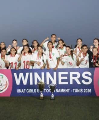 Girls soccer team posing with trophies after winning, holding a banner that reads WINNERS at a stadium during sunset.