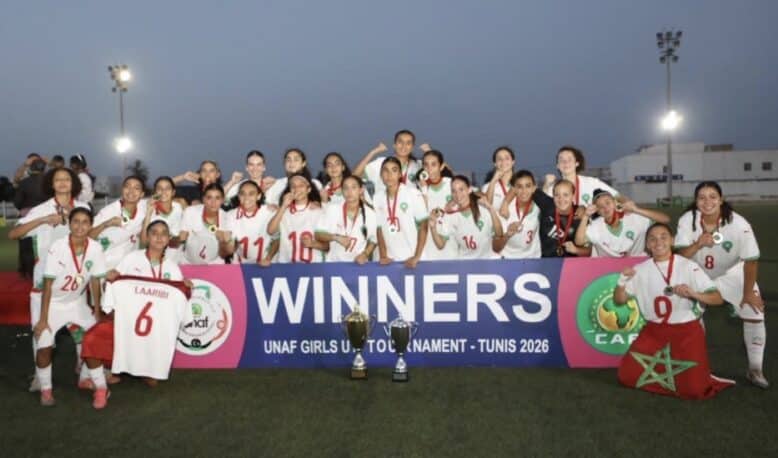 Girls soccer team posing with trophies after winning, holding a banner that reads WINNERS at a stadium during sunset.