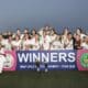 Girls soccer team posing with trophies after winning, holding a banner that reads WINNERS at a stadium during sunset.