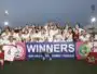 Girls soccer team posing with trophies after winning, holding a banner that reads WINNERS at a stadium during sunset.