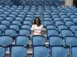 Smiling woman sitting alone in a mostly empty blue stadium seating area on a sunny day.