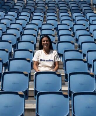 Smiling woman sitting alone in a mostly empty blue stadium seating area on a sunny day.