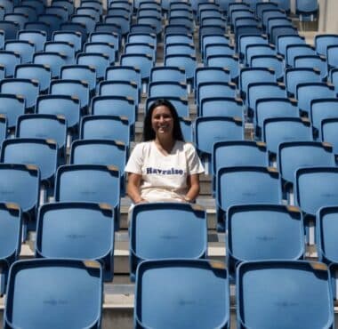 Smiling woman sitting alone in a mostly empty blue stadium seating area on a sunny day.