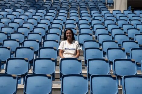 Smiling woman sitting alone in a mostly empty blue stadium seating area on a sunny day.
